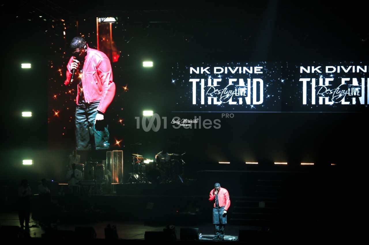 Un chanteur en veste rouge se produit sur scène avec un écran géant affichant son image et des lumières de concert.