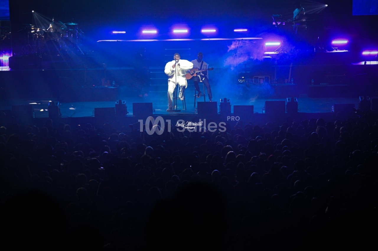 Un chanteur sur scène, éclairé par des projecteurs bleus, devant une foule dans une salle de concert.