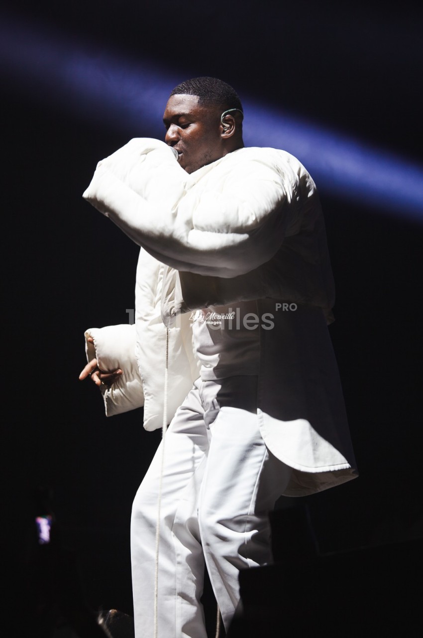 Un chanteur sur scène portant une tenue blanche ample, éclairé par un projecteur dans un environnement sombre.
