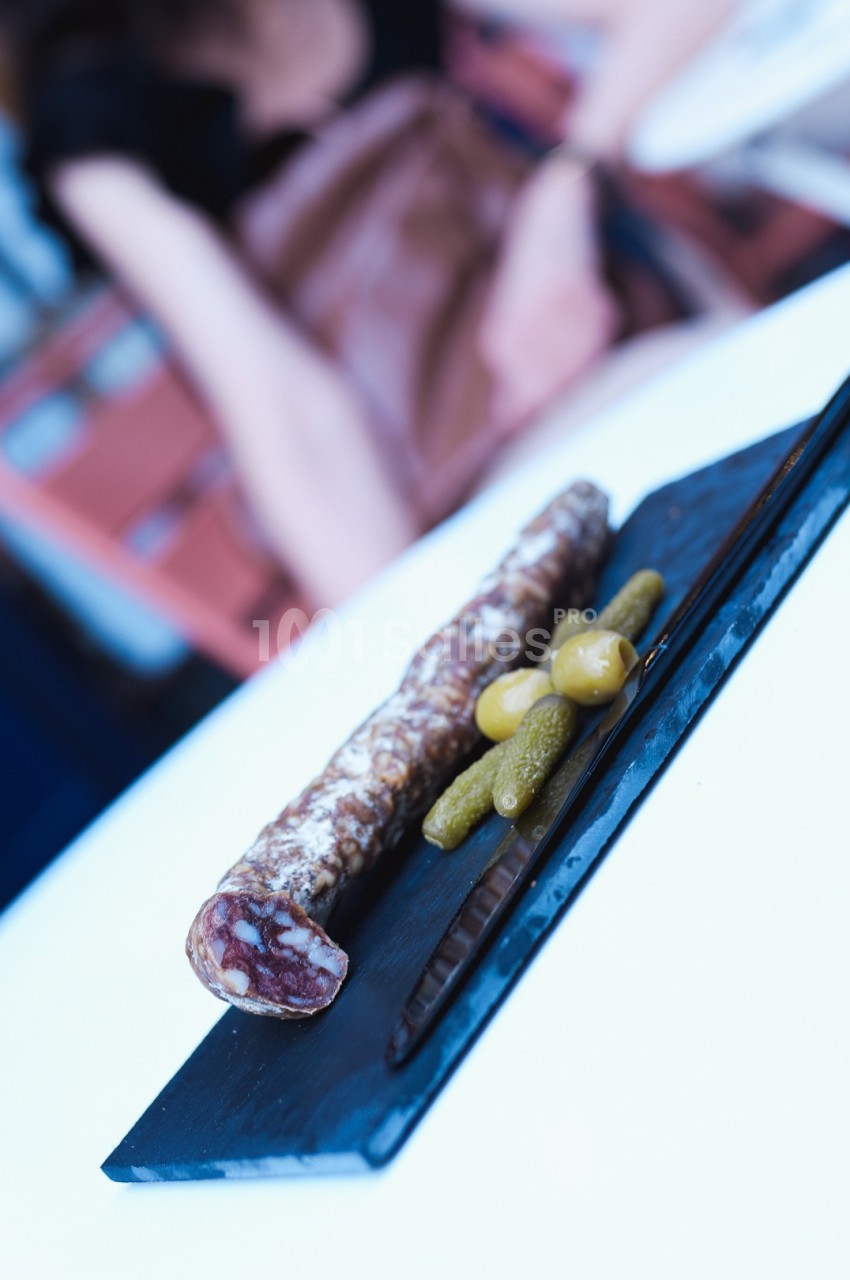 Saucisson sec tranché sur une planche avec des cornichons et des olives, posé sur une table blanche.