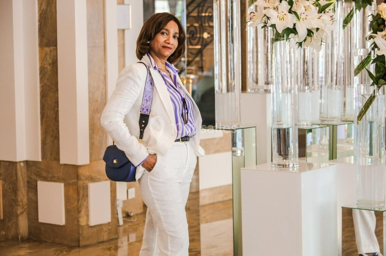 Femme debout dans un intérieur élégant, portant une tenue blanche et une chemise rayée, près de vases décoratifs.