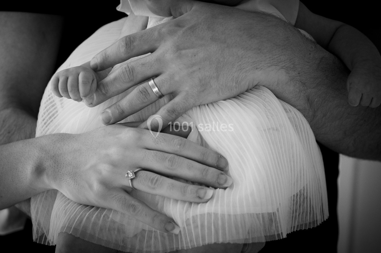 Deux mains d'adultes, l'une avec une bague, posées sur un bébé vêtu d'une robe plissée blanche.