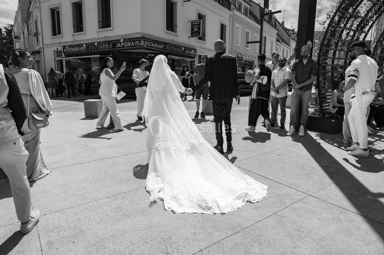 Un couple de mariés marche au centre d'une place animée, entouré de passants et de musiciens.