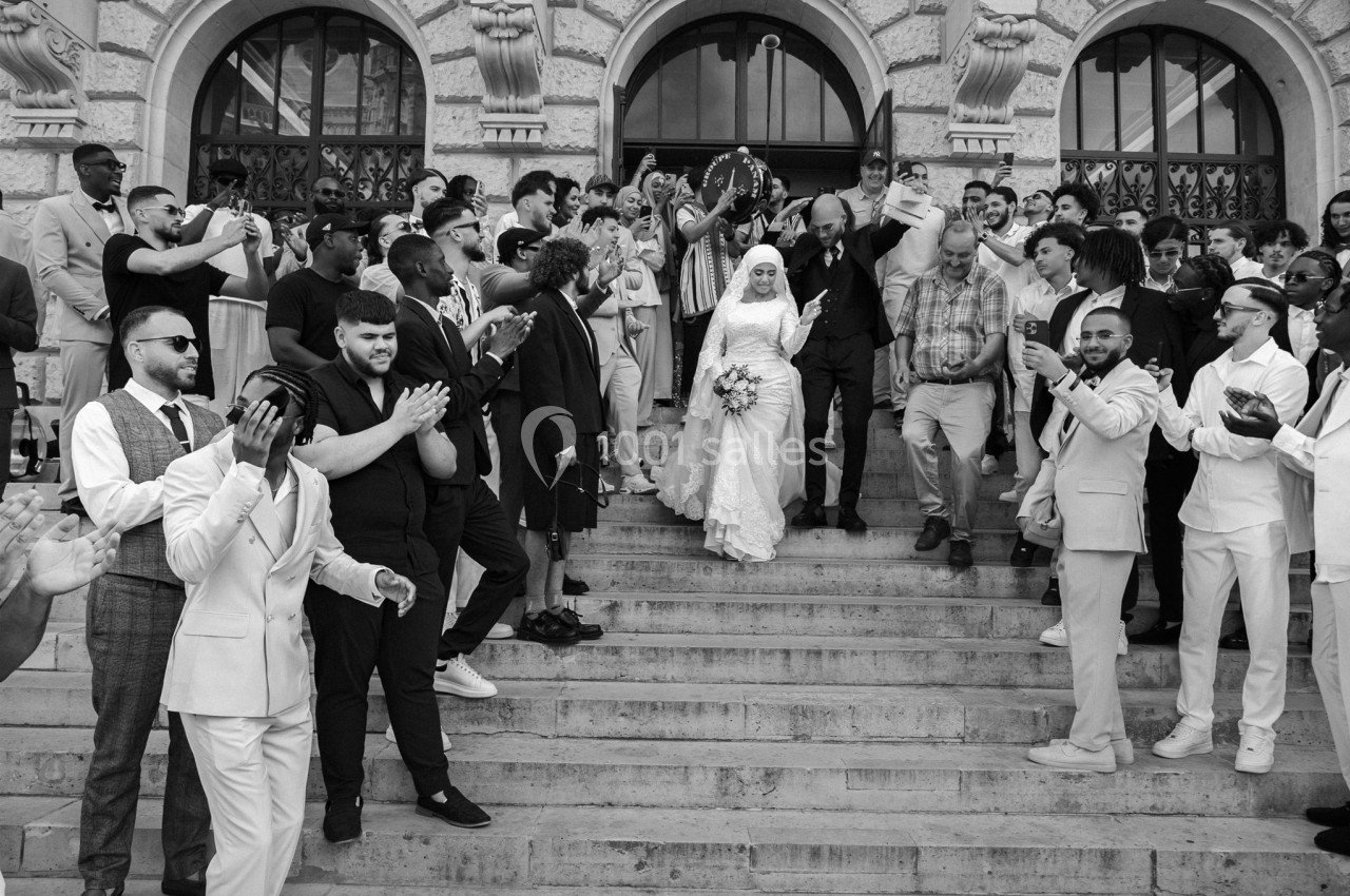 Un couple de mariés descend les marches d'un bâtiment entouré d'invités applaudissant et célébrant.