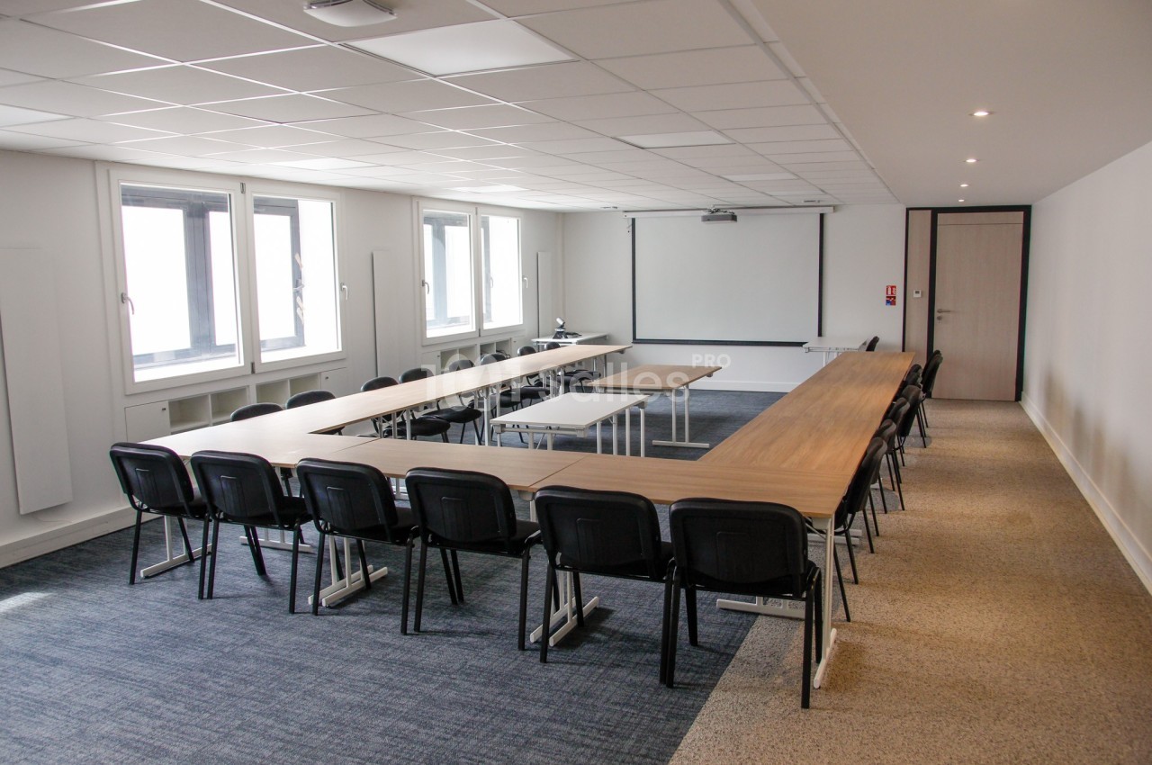 Salle de réunion lumineuse avec tables disposées en U, chaises noires, projecteur et écran blanc.