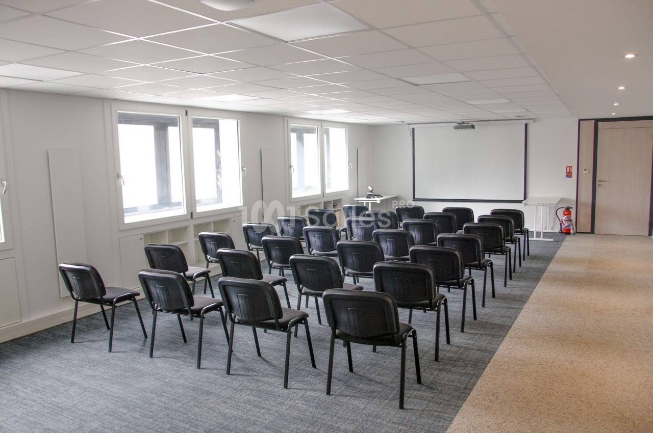Salle de réunion lumineuse avec rangées de chaises noires, écran de projection et fenêtres sur un mur latéral.