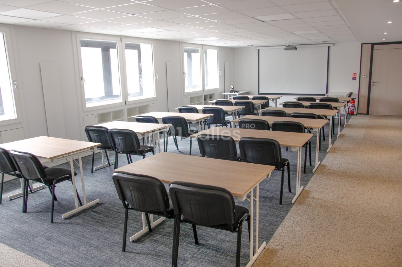 Salle de formation lumineuse avec des tables, chaises noires et un écran de projection au fond.