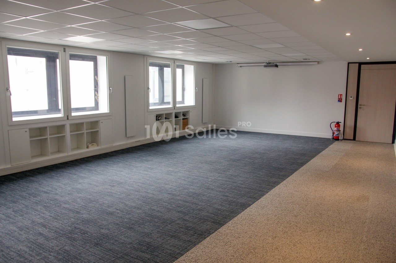 Salle vide avec moquette grise et beige, murs blancs, fenêtres vitrées, et étagères intégrées sous les fenêtres.