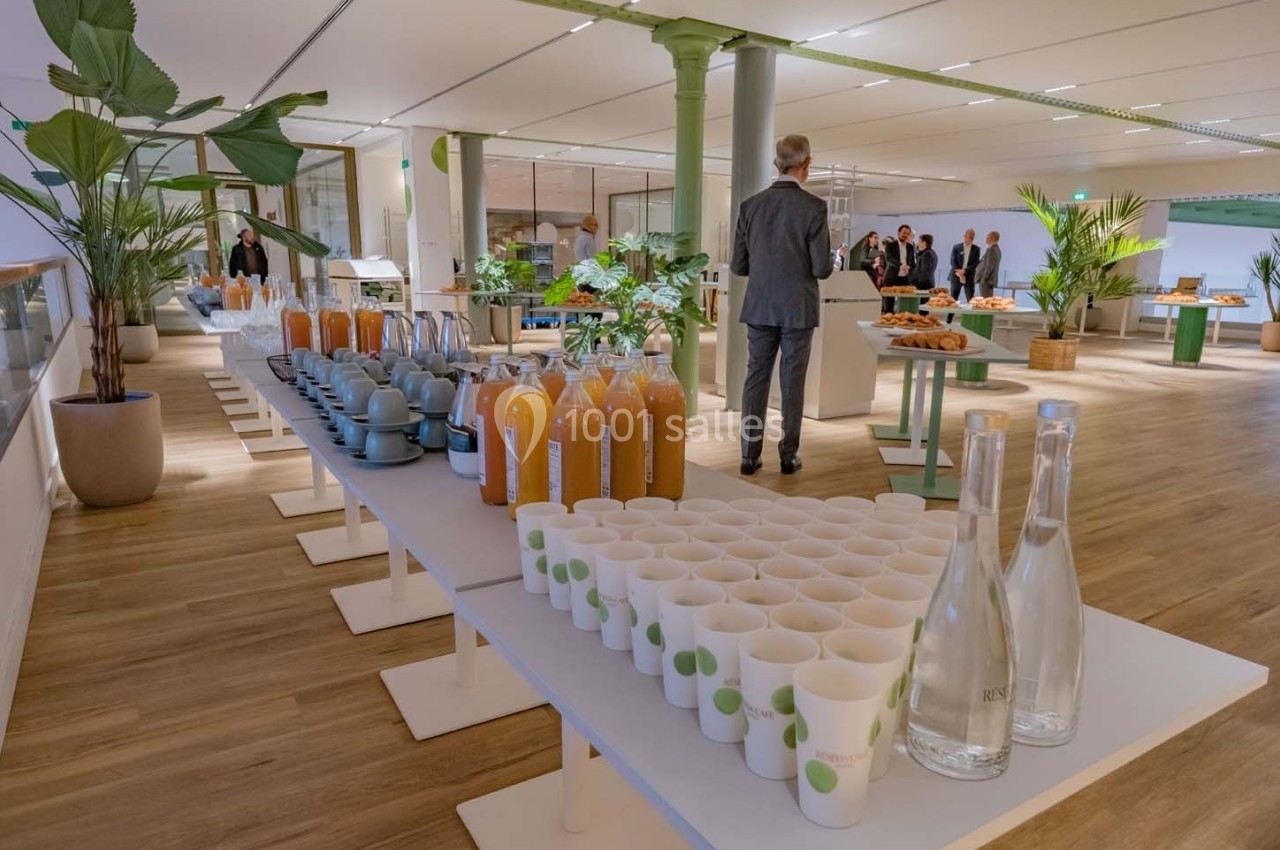 Buffet avec jus d'orange, tasses, bouteilles d'eau et plantes décoratives dans une salle lumineuse avec des invités en…