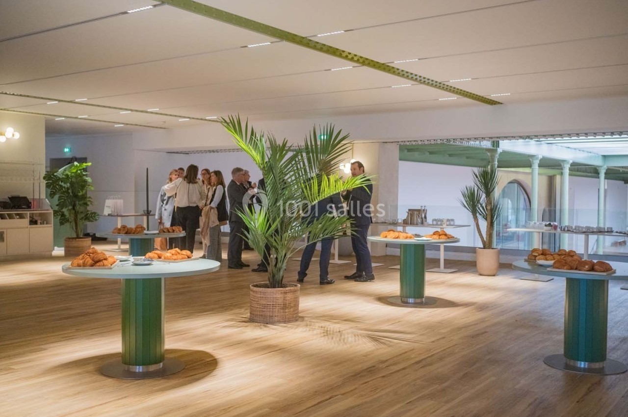 Salle lumineuse avec tables garnies de viennoiseries, plantes décoratives et groupe de personnes en arrière-plan.
