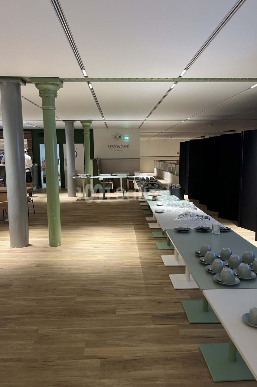 Salle lumineuse avec tables alignées, vaisselle disposée et piliers métalliques, dans un espace moderne et épuré.