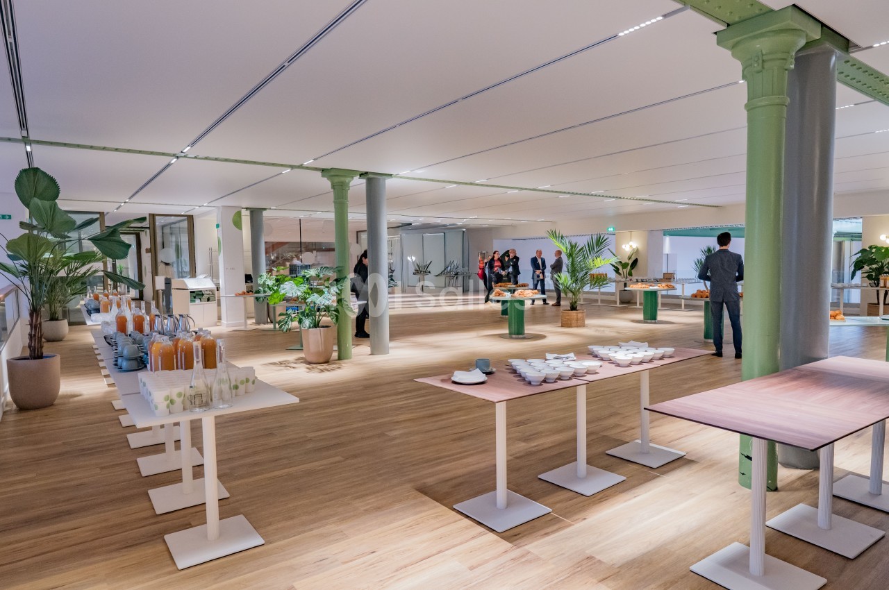 Salle lumineuse avec parquet, tables dressées pour un buffet, plantes décoratives et quelques personnes en arrière-plan.