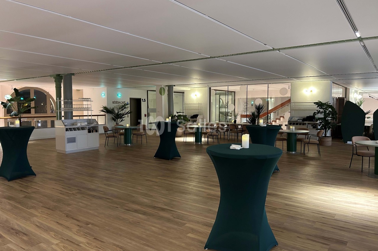 Salle lumineuse avec parquet en bois, tables hautes habillées de nappes vertes et quelques plantes décoratives.