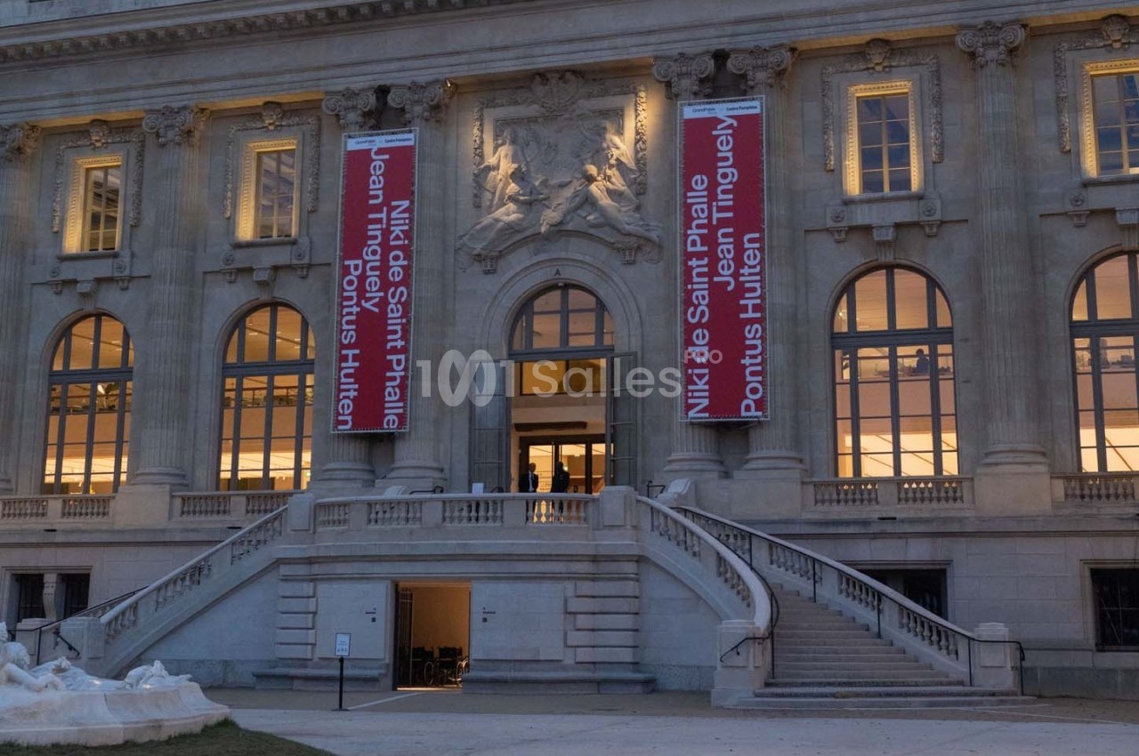 Façade d'un bâtiment historique avec des bannières rouges annonçant une exposition, vue en soirée.