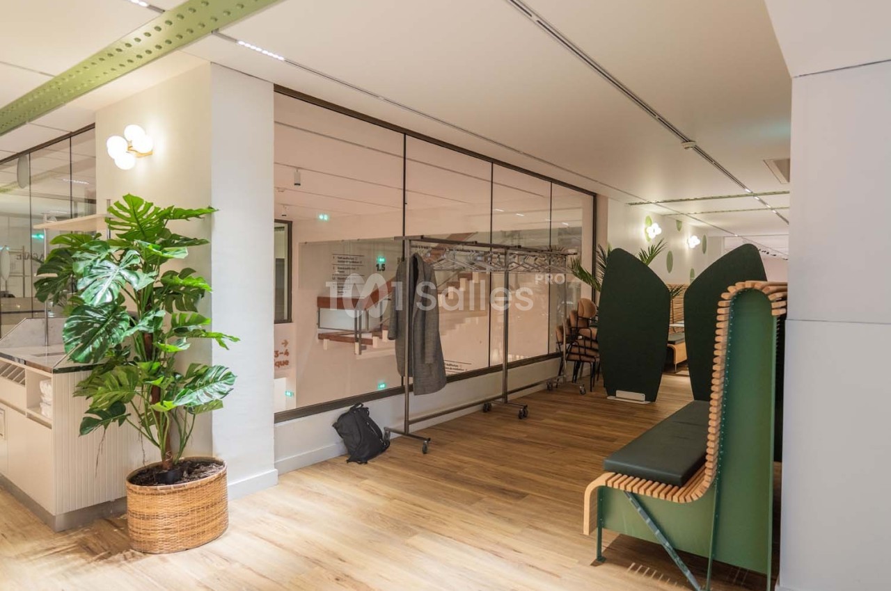 Espace intérieur lumineux avec plantes, banc en bois et métal, et cloison vitrée donnant sur un escalier.