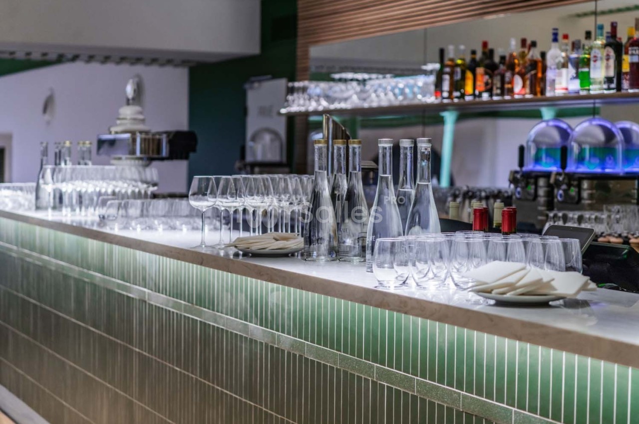 Bar moderne avec comptoir en carrelage vert, verres, bouteilles d'eau et alcools alignés sur des étagères éclairées.