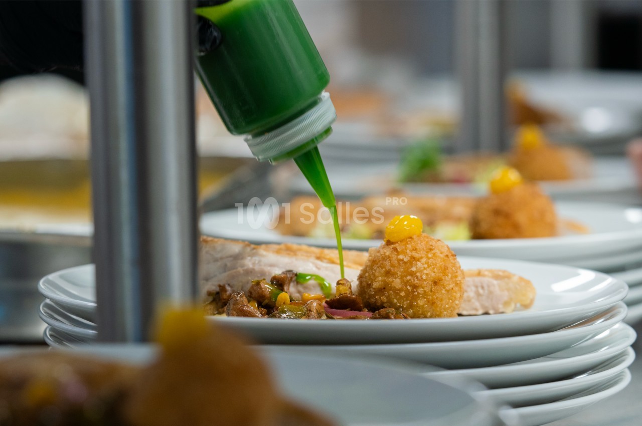Un chef verse une sauce verte sur un plat composé de boulettes croustillantes et d'accompagnements variés.