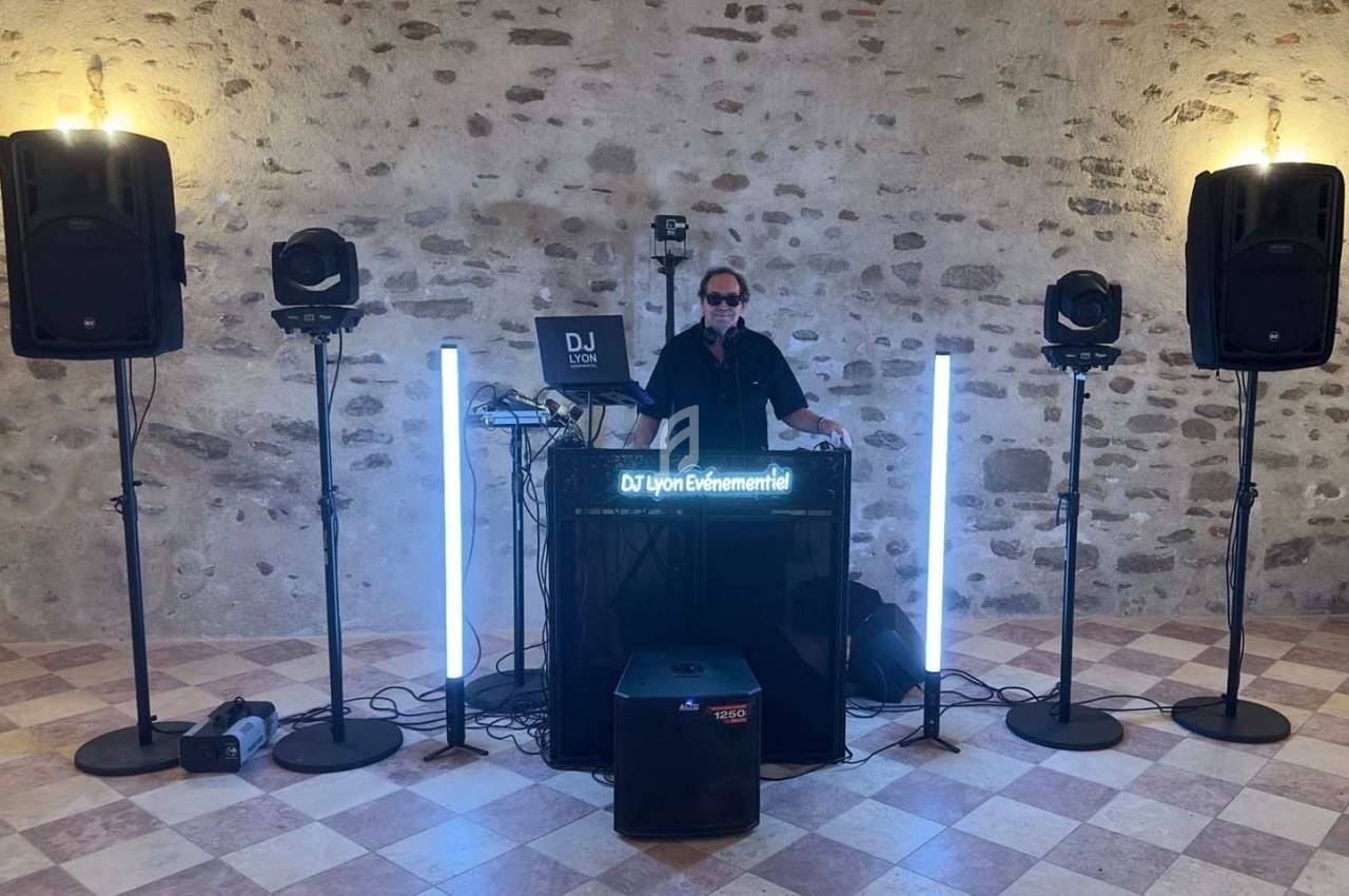 Un DJ mixe derrière une table équipée de matériel audio et lumineux dans une salle en pierre avec un sol carrelé.