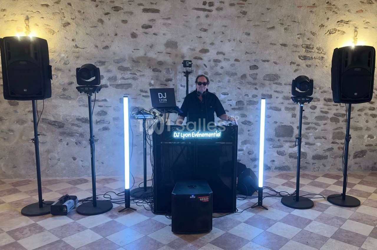 Un DJ mixe derrière une table équipée de matériel audio et lumineux dans une salle en pierre avec un sol carrelé.