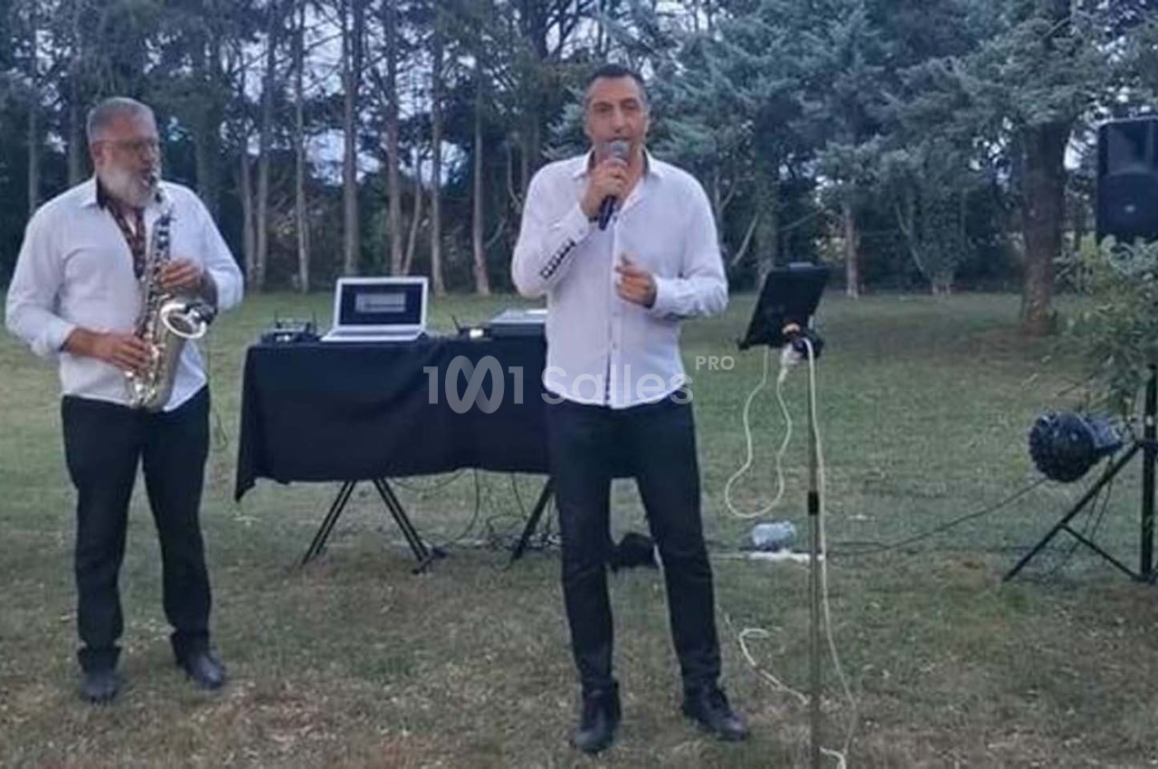 Deux musiciens jouent en plein air, l'un au saxophone et l'autre chante avec un micro devant un équipement audio.