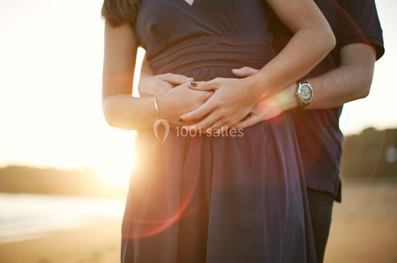 Un couple se tient enlacé sur une plage au coucher du soleil, les mains posées sur le ventre de la femme.
