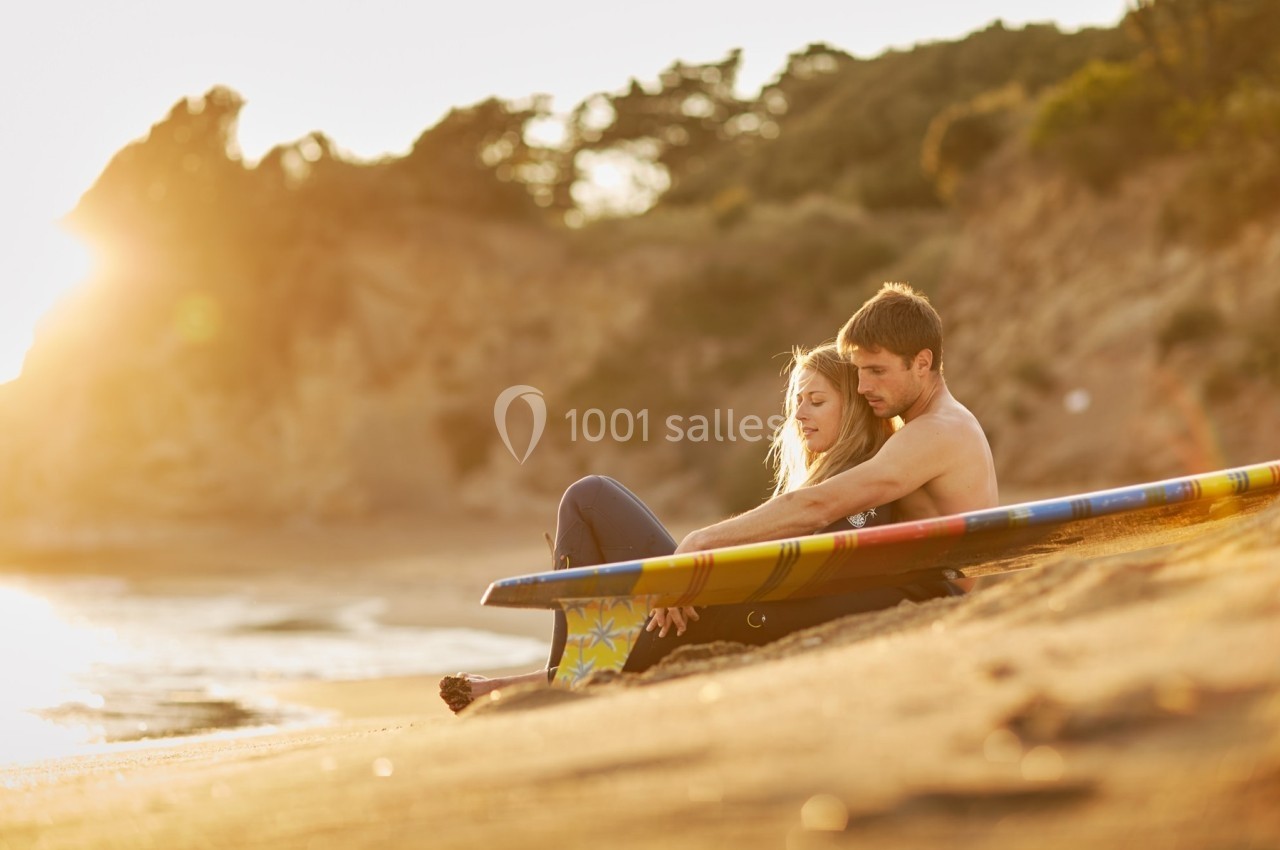 Un couple assis sur une plage au coucher du soleil, appuyé contre une planche de surf colorée.