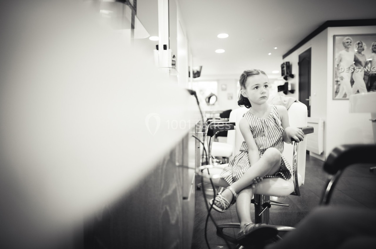 Une petite fille assise sur une chaise dans un salon de coiffure, regardant au loin avec une expression pensive.