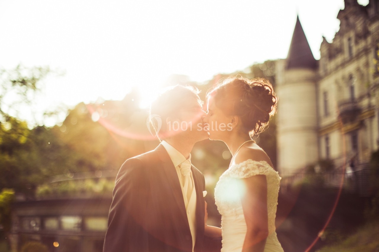 Un couple en tenue de mariage s'embrasse en extérieur, avec un château et un coucher de soleil en arrière-plan.