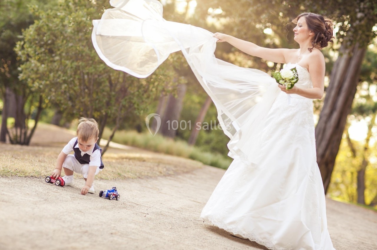 Une mariée joue avec son voile au vent près d'un enfant assis au sol, tenant des petites voitures, dans un parc.