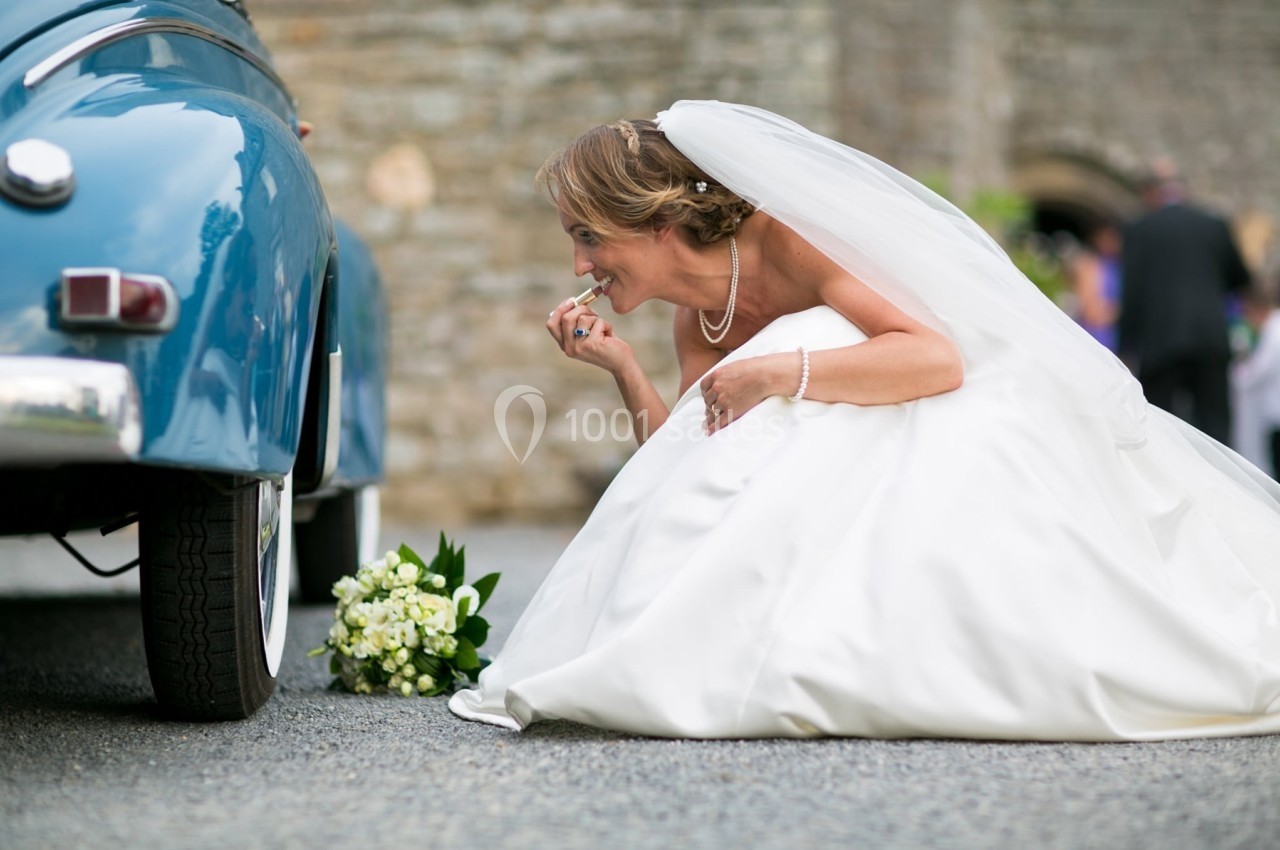 Une mariée en robe blanche, accroupie près d'une voiture bleue, ajuste son maquillage avec un bouquet posé au sol.