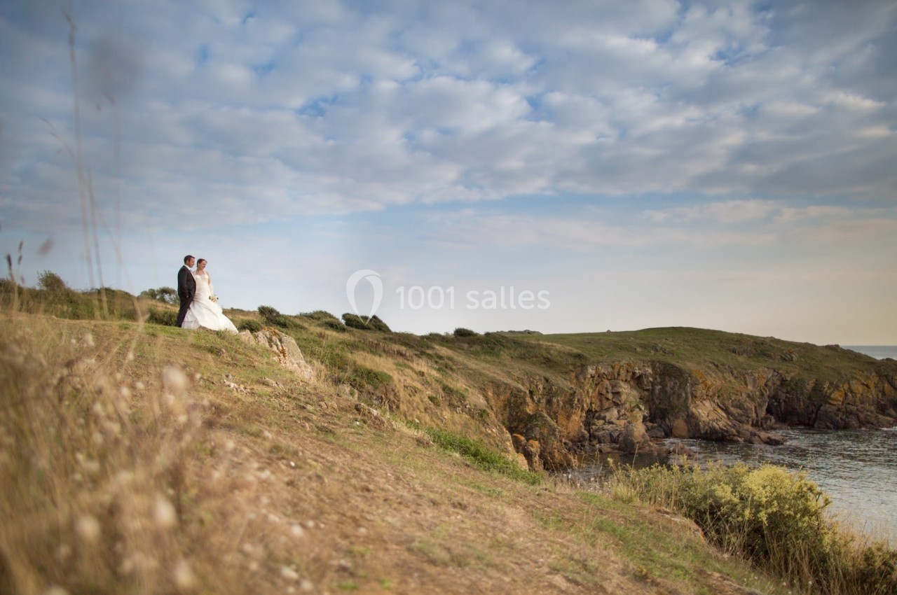 Un couple en tenue de mariage marche sur une falaise herbeuse surplombant la mer sous un ciel partiellement nuageux.
