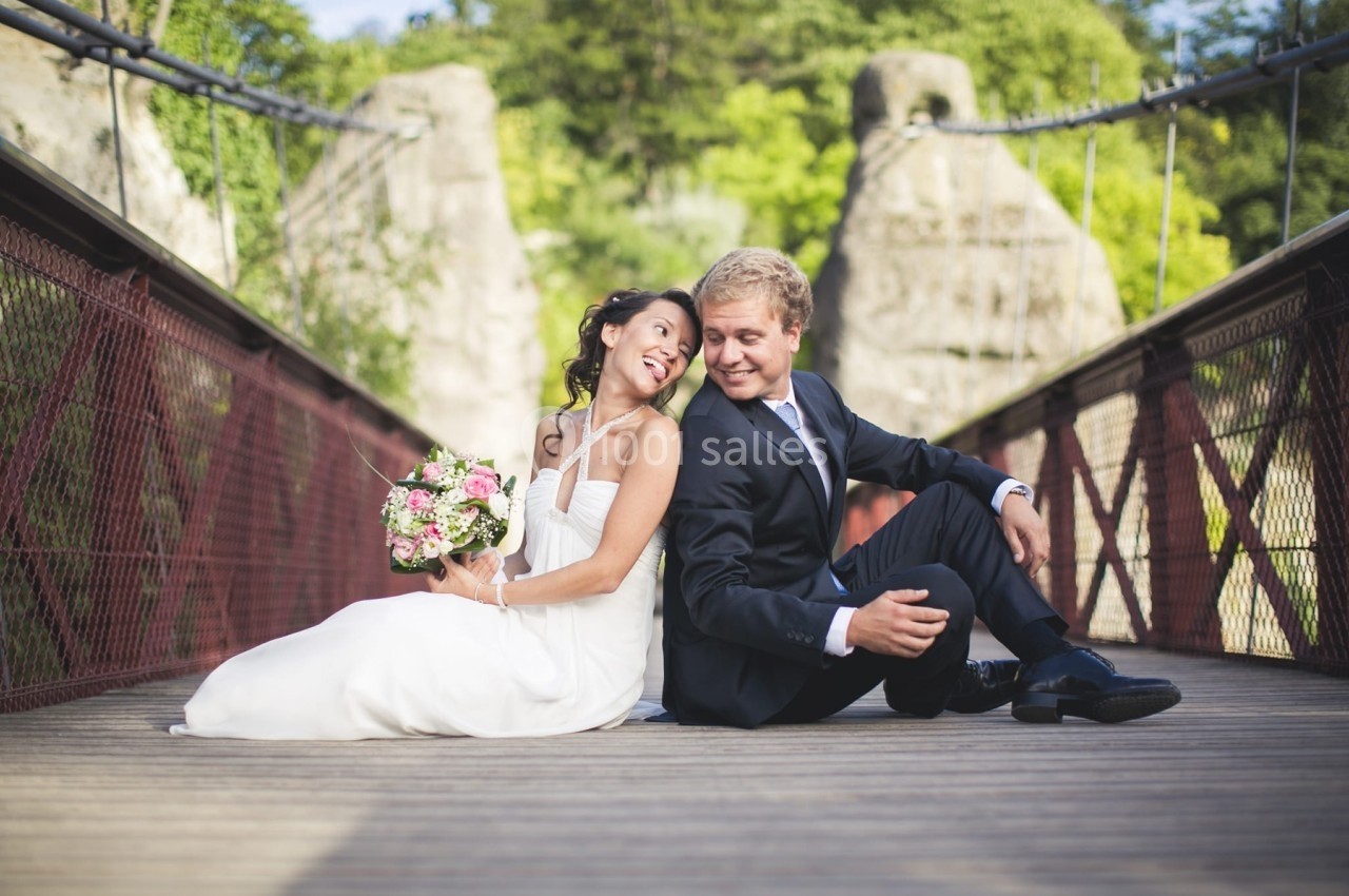 Un couple en tenue de mariage assis sur un pont en bois, entouré de verdure et de structures rocheuses.