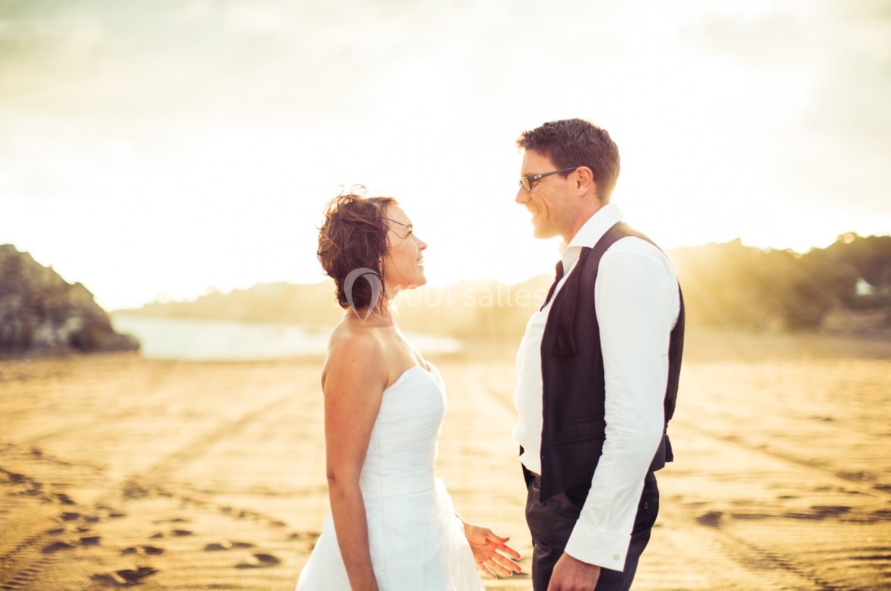 Un couple en tenue de mariage se regarde sur une plage au coucher du soleil.
