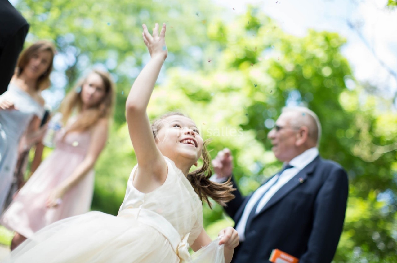 Une jeune fille en robe claire lance des confettis en l'air lors d'une célébration en extérieur, entourée de personnes.