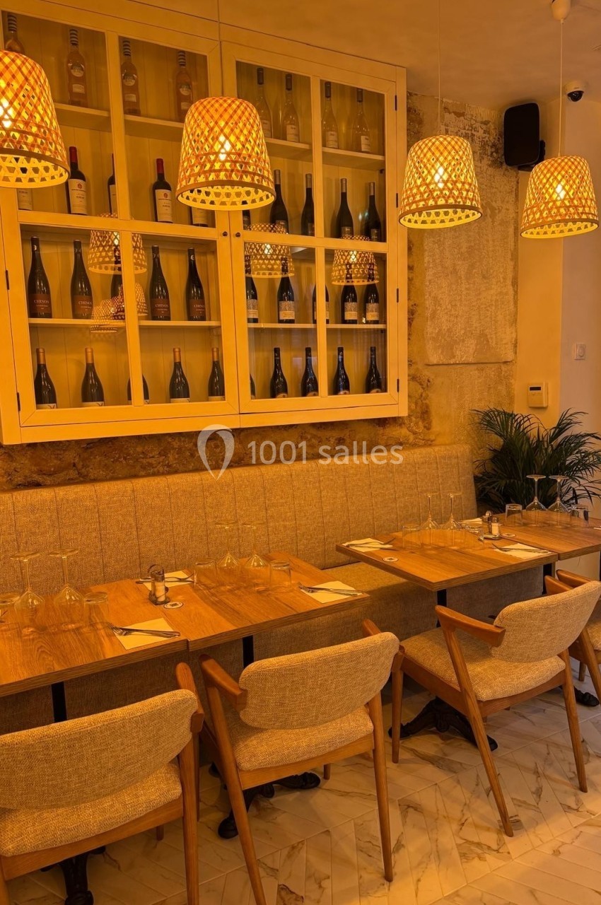 Salle de restaurant chaleureuse avec tables en bois, chaises rembourrées, suspensions lumineuses et étagère à bouteilles.