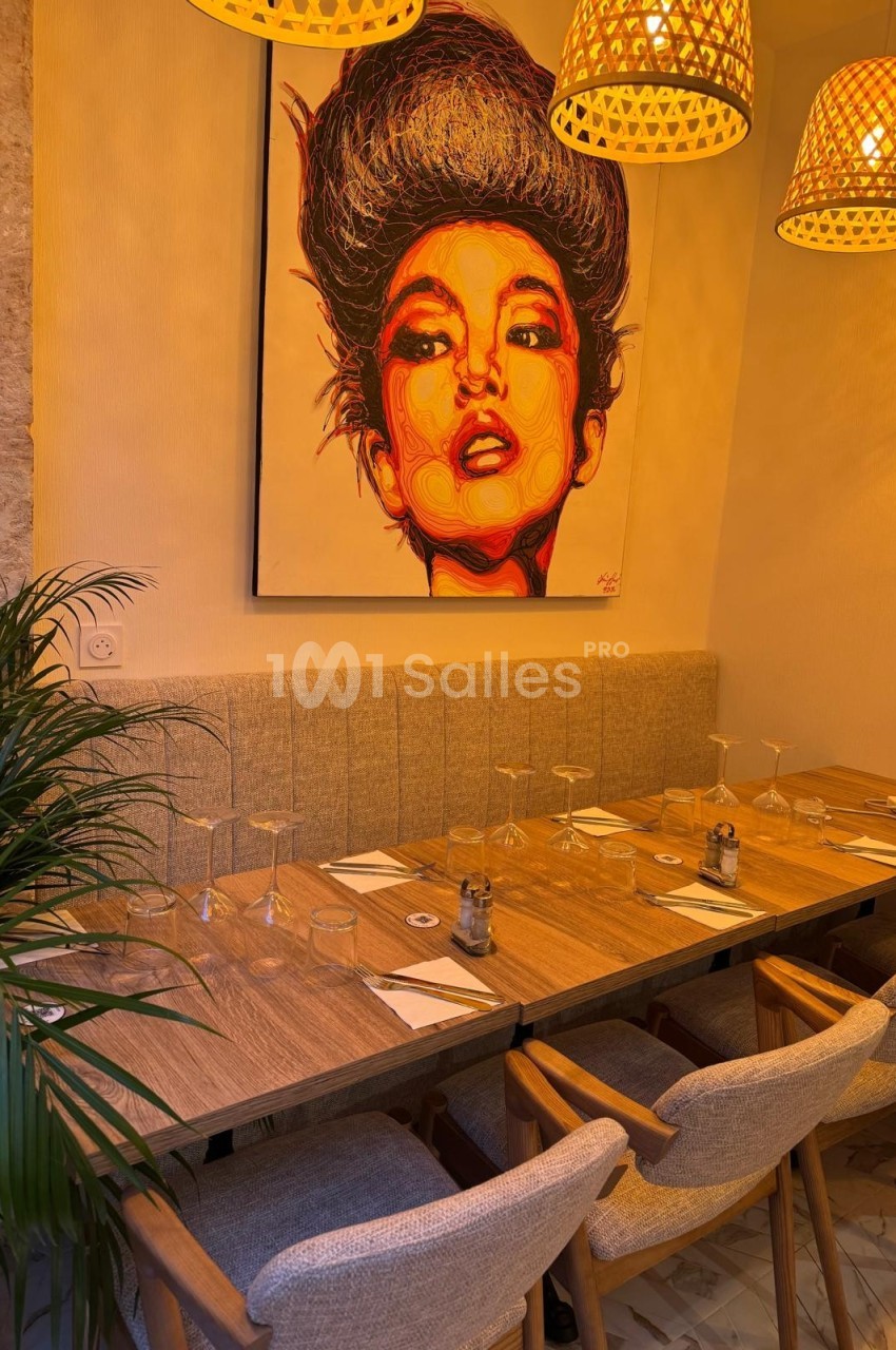 Salle de restaurant chaleureuse avec table en bois dressée, chaises rembourrées et tableau coloré au mur.