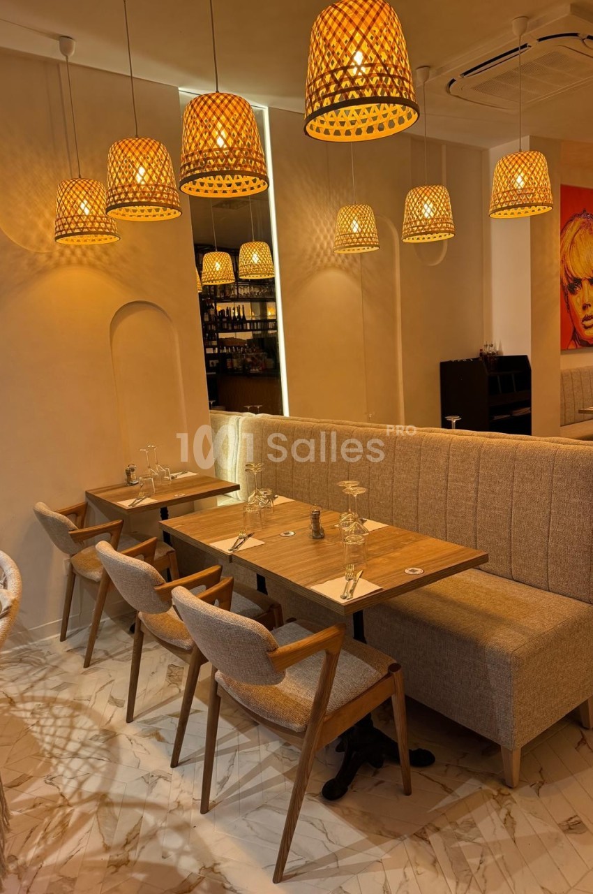 Salle de restaurant chaleureuse avec tables en bois, chaises rembourrées et suspensions en osier diffusant une lumière douce.