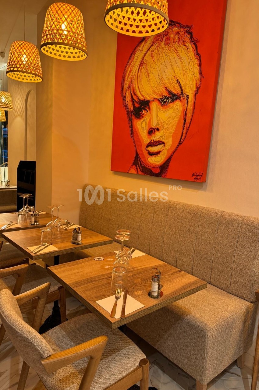 Salle de restaurant avec tables en bois, chaises rembourrées, luminaires suspendus et un tableau coloré au mur.