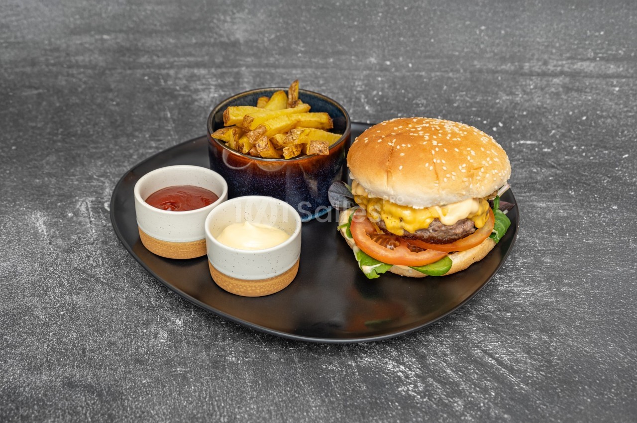 Assiette contenant un burger au fromage, des frites dans un bol et deux sauces en ramequins sur une table grise.