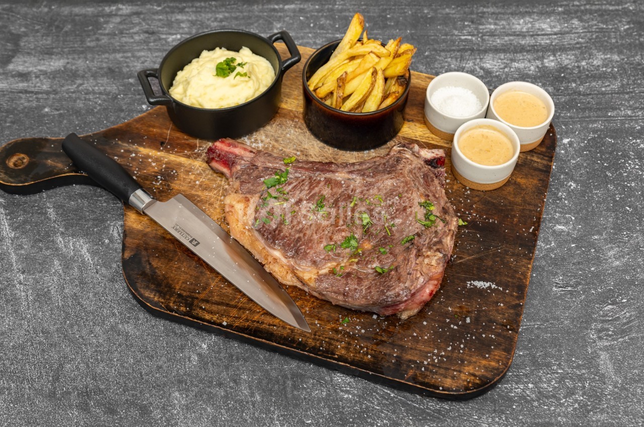 Côte de bœuf grillée servie sur une planche avec purée, frites, sauces et couteau.
