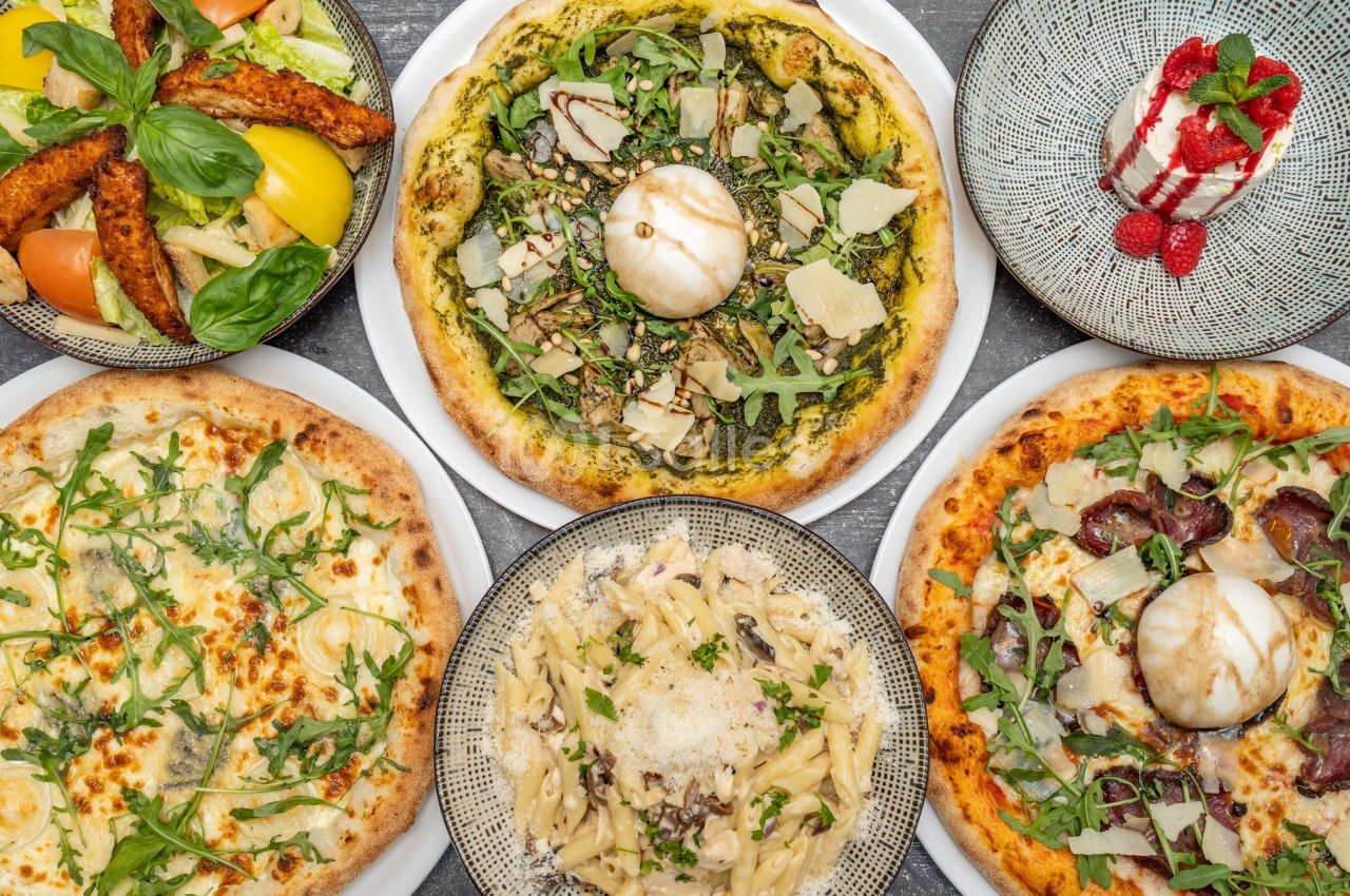 Assortiment de plats italiens comprenant pizzas, pâtes, salade et dessert sur une table grise.