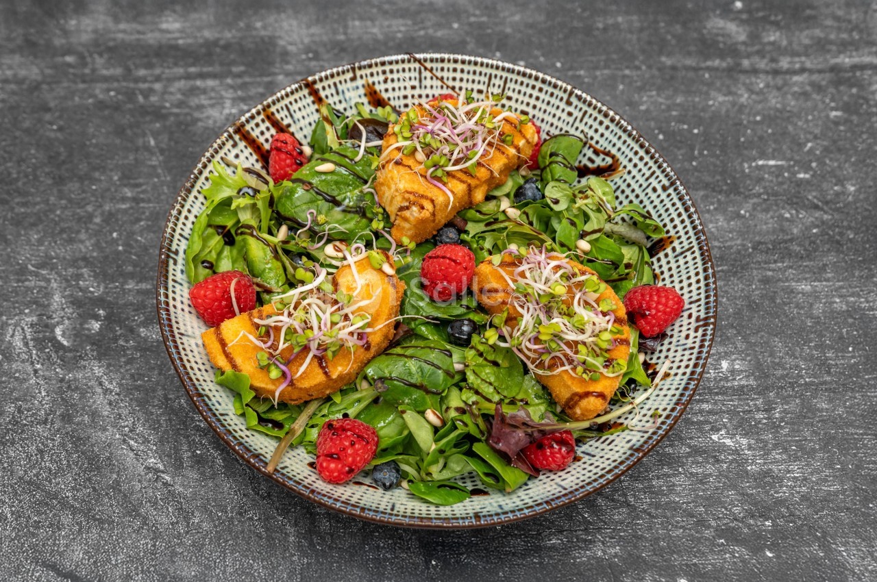 Assiette de salade verte garnie de framboises, pousses germées et rouleaux croustillants sur une table sombre.
