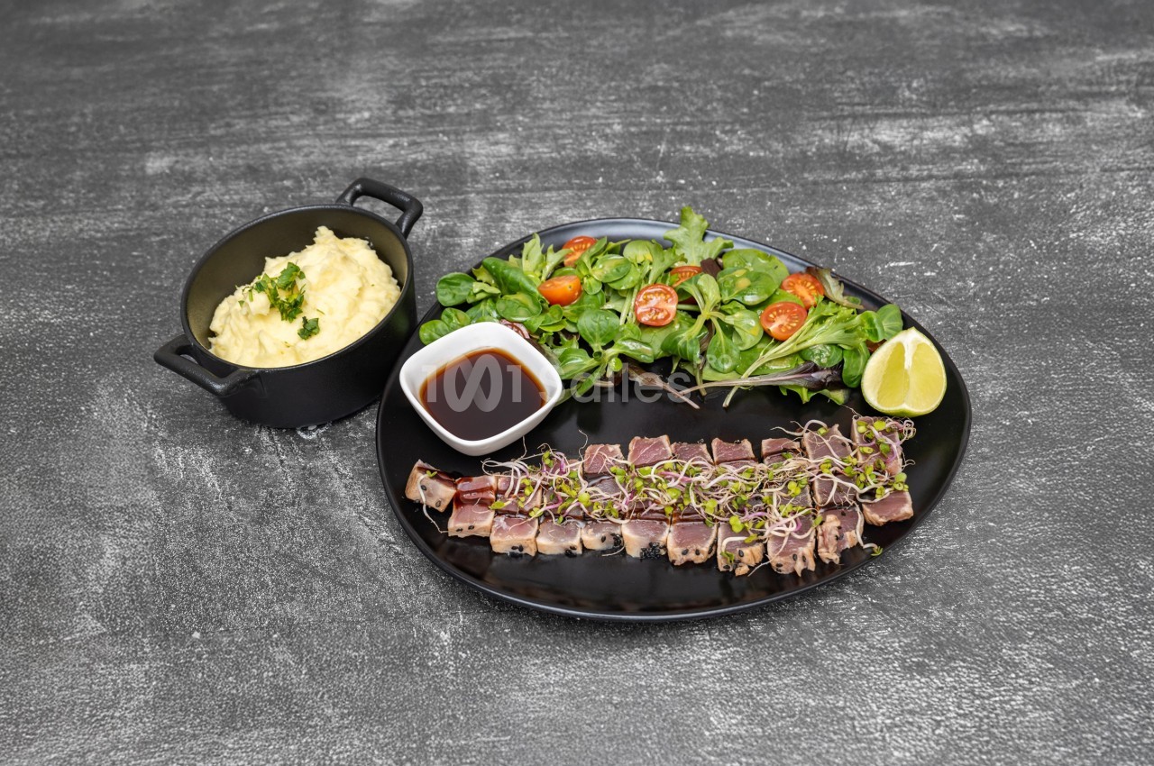 Assiette avec tataki de thon, salade verte, sauce soja, citron vert et purée servie dans une petite cocotte noire.