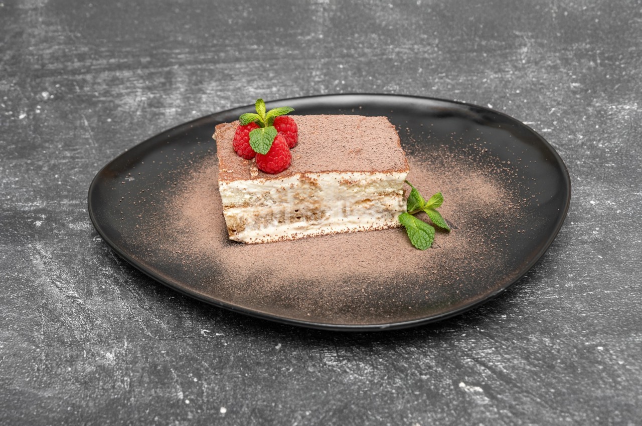 Part de tiramisu sur une assiette noire, décorée de framboises, feuilles de menthe et saupoudrée de cacao.