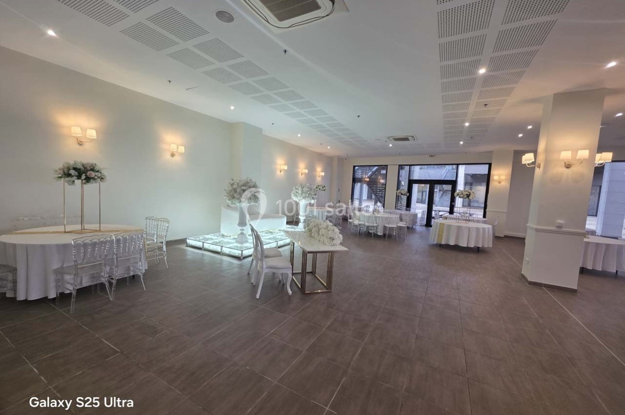 Salle de réception lumineuse avec tables rondes décorées de fleurs blanches et un sol en carrelage marron.