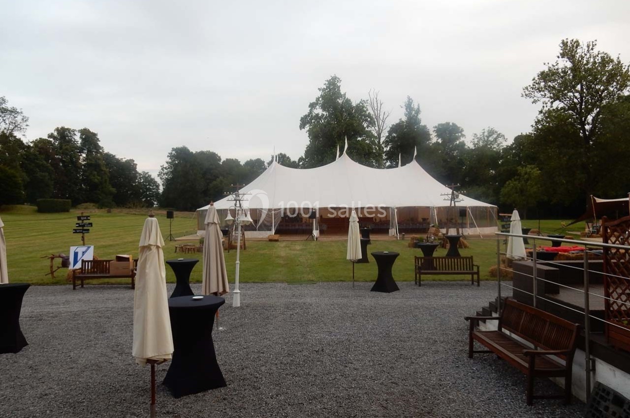 Grande tente blanche installée sur une pelouse entourée d'arbres, avec des tables et parasols disposés à l'extérieur.