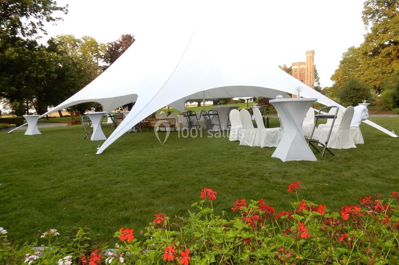Tente blanche installée sur une pelouse avec des tables et chaises, entourée de fleurs et d'arbres.