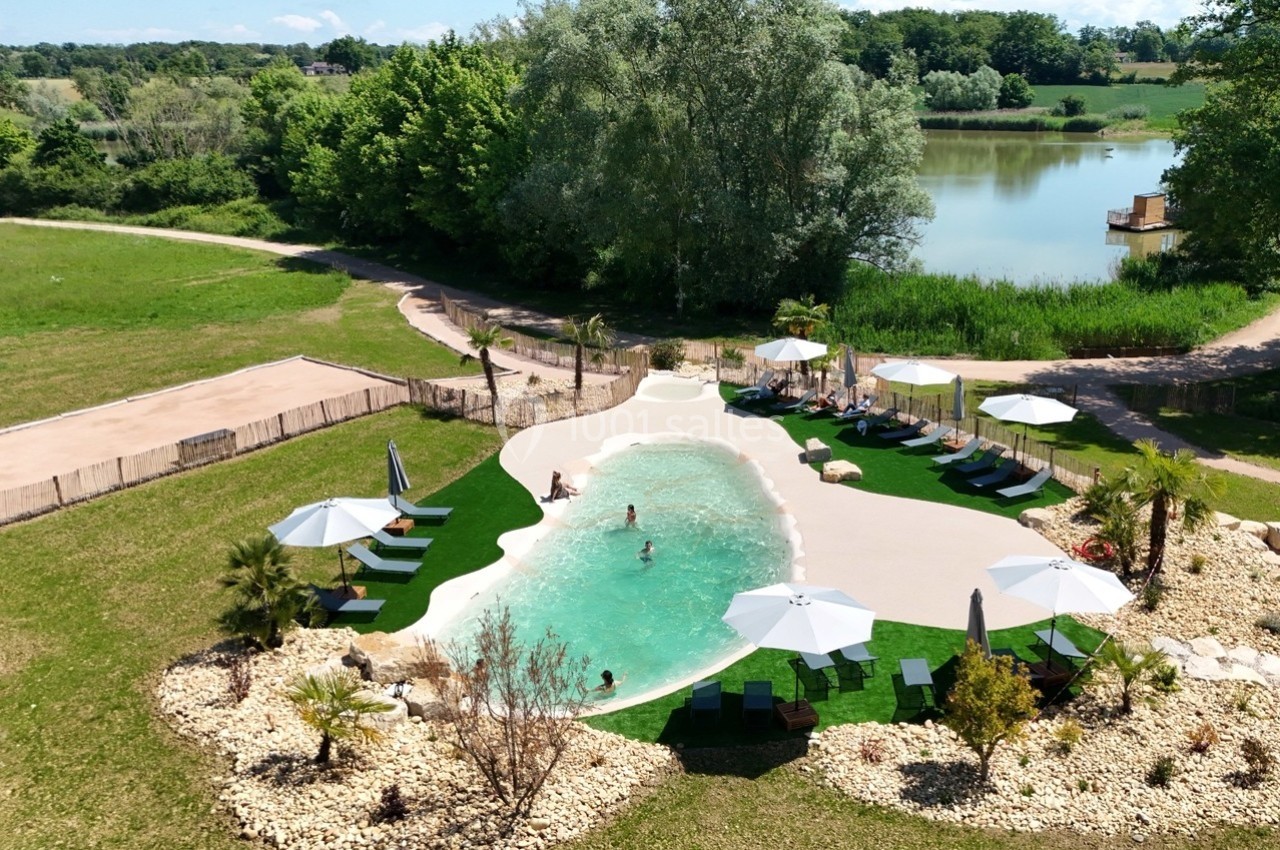 Vue aérienne d'une piscine naturelle entourée de végétation, avec transats et parasols disposés autour.