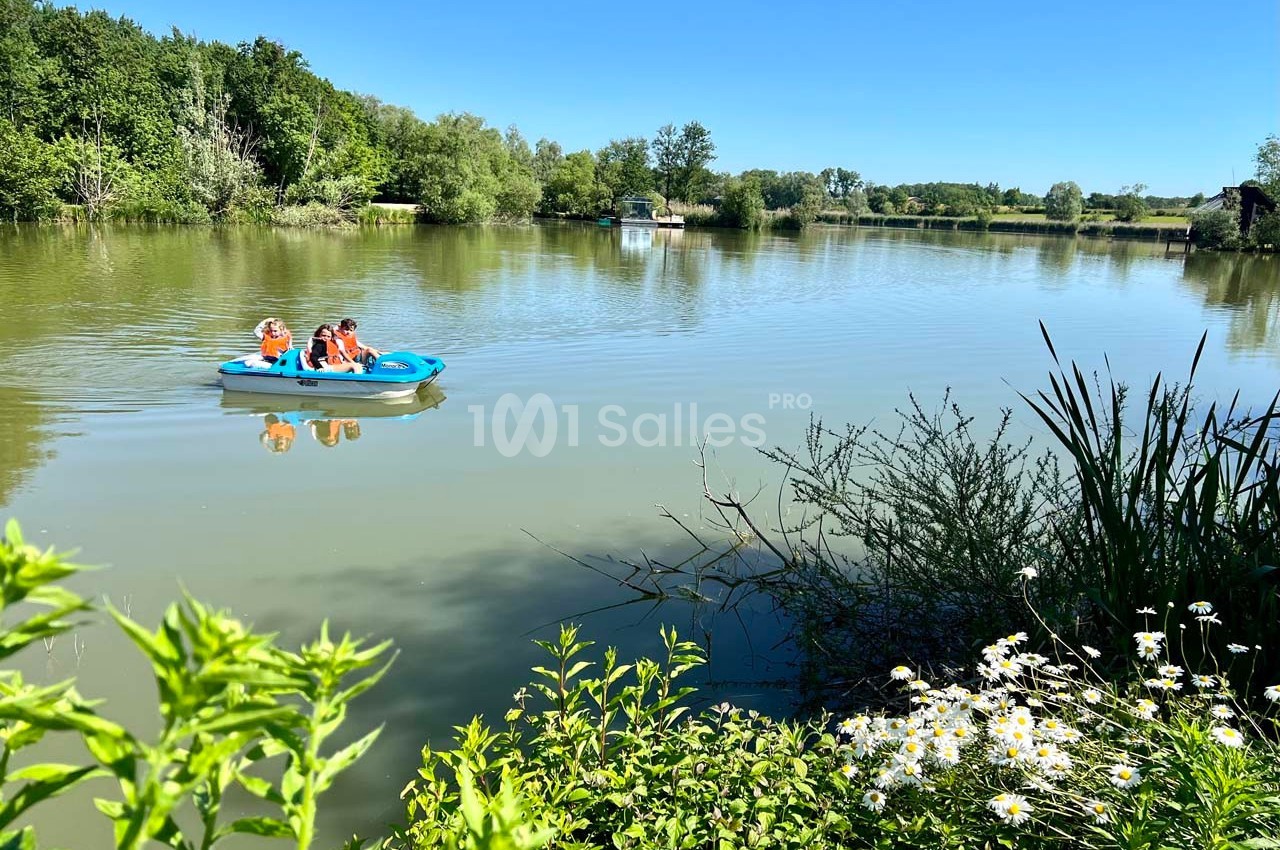 Deux personnes pagayant sur un pédalo bleu au milieu d'un lac entouré de végétation sous un ciel dégagé.