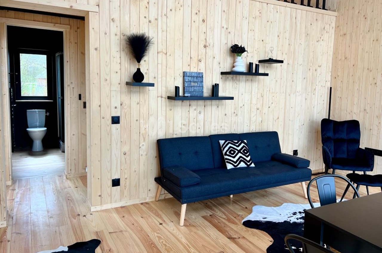 Salon en bois clair avec canapé bleu, étagères murales noires, table, chaises et vue sur une salle de bain au fond.